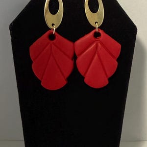 Puede incluir: Un par de pendientes rojos con forma de hoja y parte superior ovalada dorada. Los pendientes se exhiben en un soporte negro, mostrando su color vibrante y diseño único. Estos pendientes son un accesorio elegante.