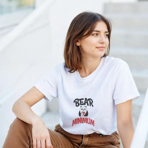 Puede incluir: Camiseta blanca con un gráfico de un panda sosteniendo un corazón y las palabras "BEAR MINIMUM" en negro y rojo. La persona lleva pantalones marrones y está sentada en escaleras blancas.