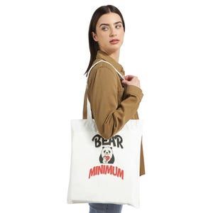 Puede incluir: Bolso tote blanco con las palabras "BEAR MINIMUM" en negro y rojo. Un oso panda sosteniendo un corazón rojo está en el centro del diseño. El bolso tiene correas blancas y se lleva al hombro.