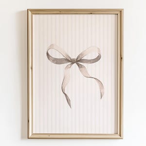May include: A framed watercolour print showing a grey bow on a striped background. The artwork is set within a gold-coloured frame, creating a classic and elegant aesthetic. The bow is the focal point, rendered in soft, muted tones.