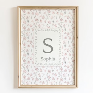 May include: Framed floral print with the name "Sophia" and the letter "S" in a gray font. The print has a light wood frame and a floral pattern with pink flowers and green leaves.