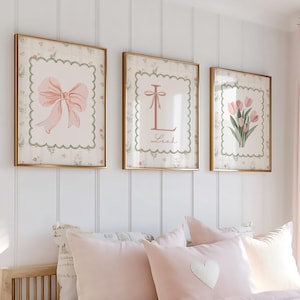 Könnte beinhalten: Drei gerahmte Wandkunstwerke mit goldenen Rahmen. Die Kunst zeigt eine rosa Schleife, den Buchstaben "L" mit einer Schleife und rosa Tulpen. Die Kunst befindet sich über einem Bett mit rosa Bettwäsche und Kissen.