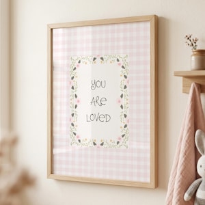 May include: A framed print with a light wood frame. The print features a pink gingham background with a white rectangle in the center. The words "you are loved" are written in black script, surrounded by a floral border. A small shelf with a vase and a towel are visible.