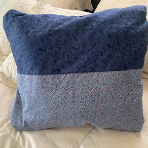 May include: A rectangular pillow with a two-tone design. The top half is navy blue with a leaf pattern, and the bottom half is light blue with a floral print. The pillow is resting on a white bed.