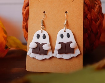 May include: A pair of Halloween-themed earrings featuring white ghost figures holding brown books. The ghosts have black eyes and are attached to silver-colored fish hook ear wires. The earrings are displayed on a brown card, with a pumpkin in the background.