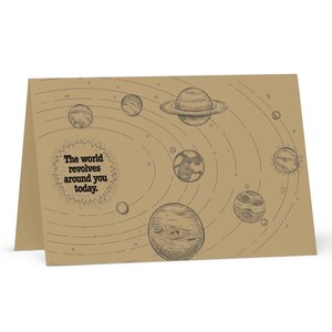 May include: A folded greeting card with a brown kraft paper finish. The card features a hand-drawn solar system design with planets and orbits. Text on the card reads, "The world revolves around you today."