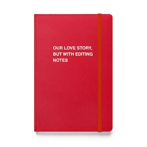 May include: A red notebook with the text "OUR LOVE STORY, BUT WITH EDITING NOTES" in white. The notebook has an orange elastic band closure running vertically along the right side.