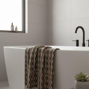 May include: A patterned bath towel draped over the edge of a white bathtub. The towel features a geometric design in shades of brown, green, and red. A small succulent in a gray pot sits on a wooden side table next to the tub.