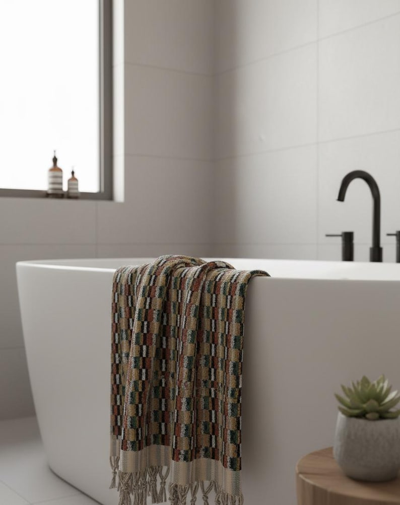 May include: A patterned bath towel draped over the edge of a white bathtub. The towel features a geometric design in shades of brown, green, and red. A small succulent in a gray pot sits on a wooden side table next to the tub.
