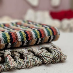 May include: Two folded towels with colorful striped patterns. The top towel has a striped pattern in shades of orange, green, yellow, and blue. The bottom towel has a fringe of knotted tassels in various colors. The towels are stacked on top of each other.