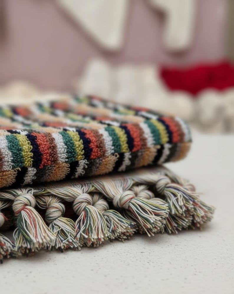 May include: Two folded towels with colorful striped patterns. The top towel has a striped pattern in shades of orange, green, yellow, and blue. The bottom towel has a fringe of knotted tassels in various colors. The towels are stacked on top of each other.