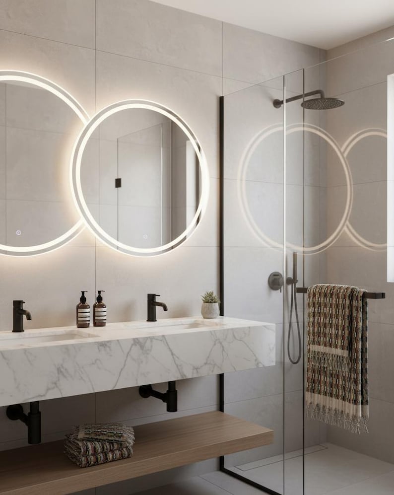 May include: Modern bathroom with two illuminated circular mirrors above a marble countertop with black faucets. A glass shower enclosure with a rain shower head and a towel rack with a patterned towel are visible. A wooden shelf holds folded towels.