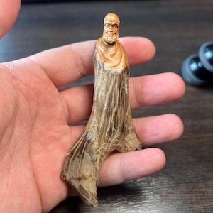Peut inclure: Une figurine en bois sculptée à la main représentant un homme barbu en robe. La figurine est brun clair avec des détails de grain de bois plus foncés, et la base est de forme naturelle et organique. La sculpture est tenue dans une main.