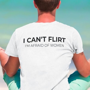 Può includere: Una t-shirt bianca a maniche corte con una scritta nera sul retro che recita I CAN'T FLIRT I'M AFRAID OF WOMEN. Il capo è realizzato in tessuto semplice e presenta un classico girocollo, offrendo un design essenziale e lineare.