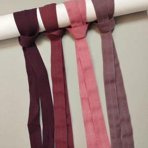 Puede incluir: Cuatro cintas de tela en tonos burdeos, rojos y rosas están anudadas alrededor de un objeto cilíndrico blanco. Las cintas cuelgan, mostrando sus variaciones de color y texturas. El fondo es gris neutro.