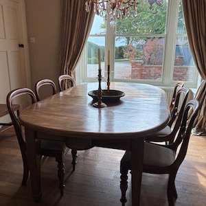 May include: A large oval wooden dining table with a smooth brown finish and turned legs, surrounded by six matching wooden chairs with upholstered seats. Two tall, thin metal candlesticks and a dark bowl sit in the centre of the table.