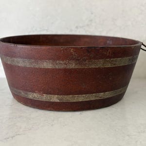 May include: A round, deep, reddish-brown wooden bowl featuring two horizontal, weathered silver-toned bands encircling the exterior. The bowl has a textured, worn finish and includes two small, thin metal ring handles attached to opposite sides.