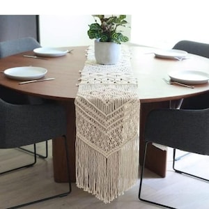 May include: A circular wooden dining table with four grey upholstered chairs. A cream macrame table runner with fringe detail runs down the centre. White plates and cutlery are set for a meal, and a potted plant sits on the table.