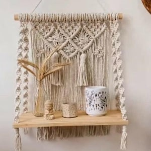 Puede incluir: Estante de pared de macramé beige con estante de madera y elementos decorativos. Incluye una pequeña estatua de Buda, un jarrón con plantas secas y una taza de cerámica estampada. El macramé cuelga de un listón de madera, añadiendo un toque bohemio.