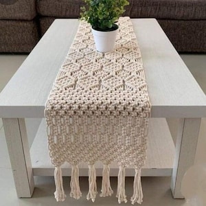 Puede incluir: Camino de mesa de macramé color crema con un patrón geométrico y flecos. Se muestra sobre una mesa de centro blanca, con una pequeña planta en maceta. El estilo general es bohemio y artesanal.