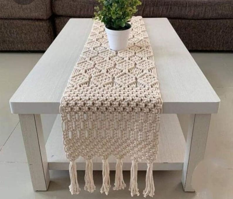 Pu&ograve; includere: Runner da tavolo in macram&egrave; color crema con motivo geometrico e frange. &Egrave; posizionato su un tavolino bianco, con una piccola pianta in vaso. L'estetica generale &egrave; boh&eacute;mien e artigianale.