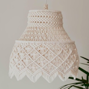 Puede incluir: Lámpara colgante de macramé color crema. La pantalla presenta intrincados patrones tejidos y un borde inferior con flecos. La luz cuelga de un cable blanco, ofreciendo un estilo de decoración bohemio o costero.