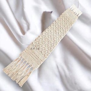 Pu&ograve; includere: Un segnalibro in macram&egrave; beige con un motivo a rombi e frange su entrambe le estremit&agrave;. Il segnalibro &egrave; realizzato in cotone intrecciato ed &egrave; esposto su uno sfondo bianco.