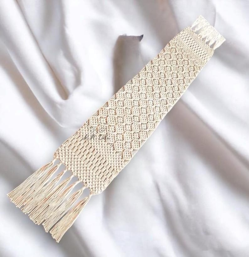 Pu&ograve; includere: Un segnalibro in macram&egrave; beige con un motivo a rombi e frange su entrambe le estremit&agrave;. Il segnalibro &egrave; realizzato in cotone intrecciato ed &egrave; esposto su uno sfondo bianco.