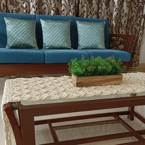 Pu&ograve; includere: Scena di soggiorno con un divano blu e tre cuscini verde acqua. Un tavolino in legno espone un runner in macram&egrave; e una fioriera in legno con piante. Tende marroni sullo sfondo.