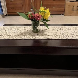 Pu&ograve; includere: Un tavolino rettangolare marrone scuro con un runner in macram&egrave; color crema. Un vaso di vetro con un bouquet di fiori gialli e rossi &egrave; posto sul runner. Il tavolo ha un ripiano inferiore. Il tavolo &egrave; lungo circa 120 cm.