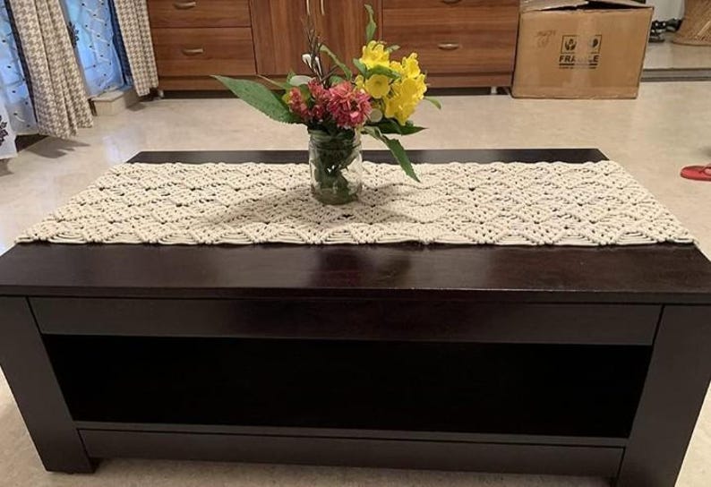 Pu&ograve; includere: Un tavolino rettangolare marrone scuro con un runner in macram&egrave; color crema. Un vaso di vetro con un bouquet di fiori gialli e rossi &egrave; posto sul runner. Il tavolo ha un ripiano inferiore. Il tavolo &egrave; lungo circa 120 cm.