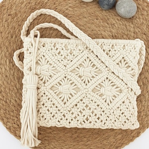 May include: Cream macrame shoulder bag with a braided strap and tassel. The bag has a geometric floral pattern and is displayed on a woven mat with three small stones.