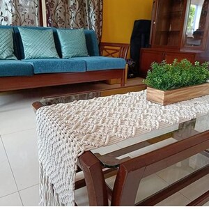 Pu&ograve; includere: Scena di soggiorno con un divano verde acqua, tre cuscini decorativi, un tavolino in legno con un runner in macram&egrave; e una fioriera in legno con piante verdi. L'ambiente &egrave; caldo e accogliente.