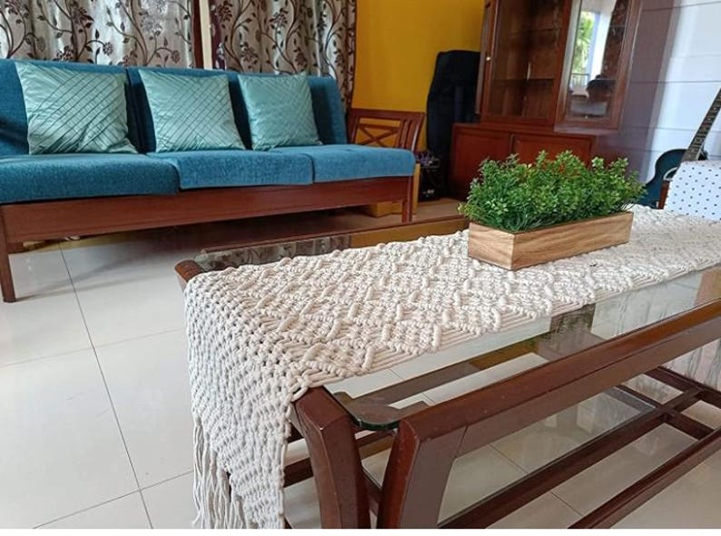 Pu&ograve; includere: Scena di soggiorno con un divano verde acqua, tre cuscini decorativi, un tavolino in legno con un runner in macram&egrave; e una fioriera in legno con piante verdi. L'ambiente &egrave; caldo e accogliente.