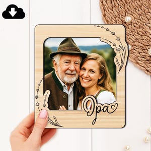Op de afbeelding: Houten fotolijst met een foto van een oudere man en een jongere vrouw. De lijst heeft een decoratief bloemmotief en het woord "Opa" in script. De foto is tegen een vage buitenachtergrond geplaatst.