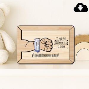 May include: Wooden plaque with a drawing of a baby's hand holding a hospital bracelet. The plaque includes the date, time, weight, and length of the baby. The text "WILLKOMMEN KLEINES WUNDER!" is at the bottom.