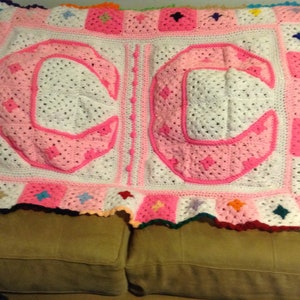 Puede incluir: Una manta de bebé de ganchillo con las letras "CC" en rosa sobre un fondo blanco. La manta tiene un borde colorido con cuadrados de diferentes colores.