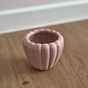 Könnte beinhalten: Ein kleiner, hellrosa Keramikübertopf mit einem abgerundeten, gewellten Außendesign. Der Topf hat eine glatte, glänzende Oberfläche und eine weite, offene kreisförmige Öffnung, wobei vertikale Rillen ein blütenblattartiges Aussehen erzeugen.