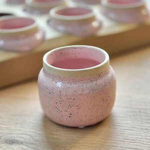 Könnte beinhalten: Eine kleine, runde Keramiktasse mit hellrosa Glasur und dunklen Sprenkeln. Der Rand der Tasse ist mit einem kontrastierenden, unglasierten beigen Band versehen. Die Tasse hat eine weite Öffnung und einen abgerundeten Körper in handgefertigter Optik.