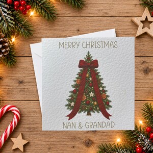 May include: A square white greeting card featuring a green Christmas tree illustration decorated with red ornaments, gold baubles, and dried orange slices. A large dark red ribbon is tied around the tree. The text reads Merry Christmas Nan & Grandad.