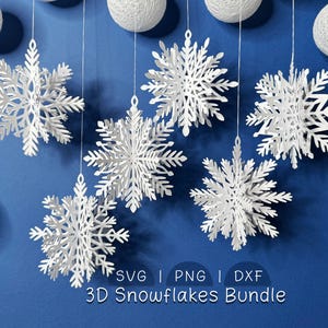 Könnte beinhalten: Weiße 3D-Papier-Schneeflocken hängen vor blauem Hintergrund, mit weißen Lichterketten und dekorativen weißen Kugeln. Der Text "SVG | PNG | DXF 3D Snowflakes Bundle" ist im Bild enthalten.