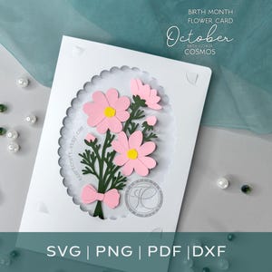 May include: A white rectangular card featuring a scalloped oval cutout displaying a layered paper craft of pink cosmos flowers with dark green stems and a small pink bow. The text reads BIRTH MONTH FLOWER CARD October COSMOS.