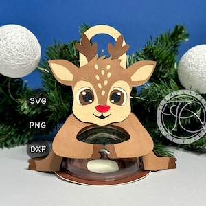 Può includere: Una renna di carta marrone con un naso rosso e grandi occhi, che tiene un contenitore trasparente. La renna ha una maniglia in alto. L'immagine include il testo "SVG", "PNG" e "DXF". Lo sfondo presenta rami di albero di Natale e palline decorative bianche.