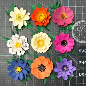 May include: Nine colorful paper flowers in various shades of white, orange, pink, blue, and purple, each with a yellow center. Green leaves are attached to the base of each flower. The flowers are arranged on a gray grid surface.