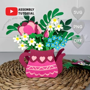 Op de afbeelding: Een papieren bloemstuk in een bordeauxrode theepotvormige vaas. Het arrangement bevat roze pioenrozen, witte madeliefjes, blauwe vergeet-mij-nietjes en groene bladeren. De vaas heeft hart- en geometrische ontwerpen. De tekst "Assembly Tutorial" en "tcartcraft.etsy.com" is inbegrepen.