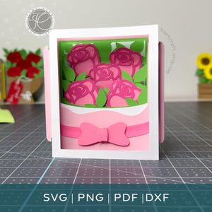 Puede incluir: Una caja de sombras de papel en capas que presenta un ramo de rosas rosadas con hojas verdes, encerrado en un marco blanco. El ramo está envuelto en papel rosa claro y acentuado con un lazo rosa. El texto SVG | PNG | PDF | DXF es visible.
