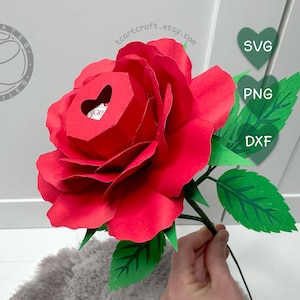 Puede incluir: Una gran rosa de papel roja con un recorte en forma de corazón en el centro. La rosa tiene hojas verdes y un tallo oscuro. La imagen también incluye el texto "SVG", "PNG" y "DXF" en gráficos verdes en forma de corazón.