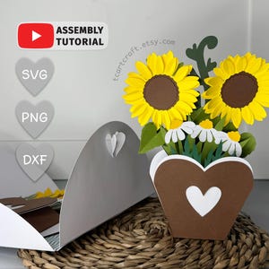 Könnte beinhalten: Ein Papierblumenarrangement mit Sonnenblumen und Gänseblümchen in einer herzförmigen Vase. Die Sonnenblumen haben gelbe Blütenblätter und braune Zentren. Die Vase ist braun mit einem weißen Herz-Ausschnitt. Der Text "ASSEMBLY TUTORIAL" ist enthalten.