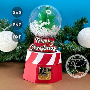 Puede incluir: Una máquina de chicles navideña roja y blanca con un globo transparente que contiene un árbol verde y acentos de copos de nieve blancos. El letrero superior dice "Merry Christmas". Adornos navideños decorativos y ramas de hoja perenne en el fondo.