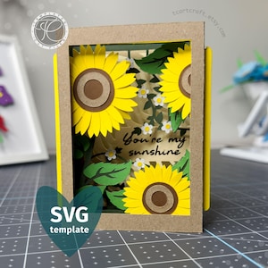 May include: A layered paper craft shadow box featuring three bright yellow sunflowers with brown centres, green leaves, and small white flowers. The centre of the box displays the black text "You're my sunshine" on a light background.